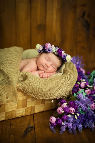 Newborn baby girl with a wreath in a wicker basket with a bouquet of purple wild flowers — Stock Photo, Image