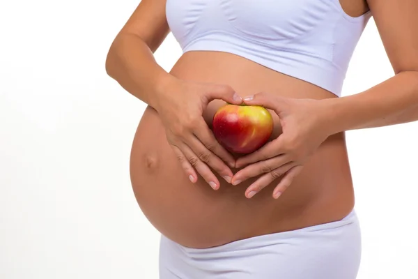 Крупним планом вагітний живіт і яблуко. Руки складені серцем. На білому тлі — стокове фото