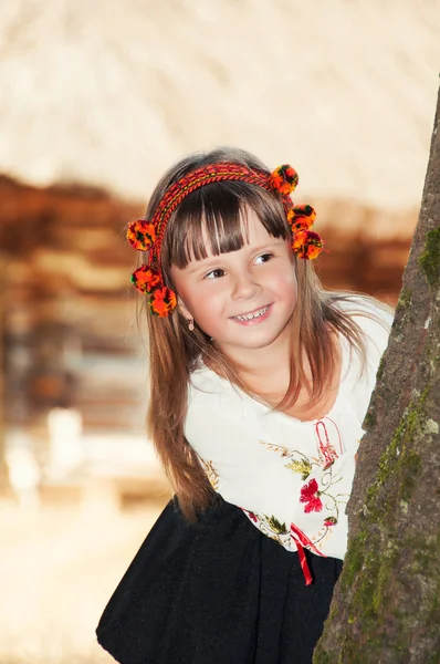 Niña sonriente con el traje nacional ucraniano Hutsul y en una corona de flores mira hacia fuera desde detrás de un árbol — Foto de Stock