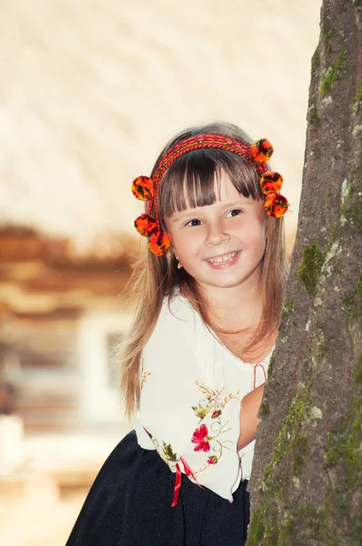 Niña sonriente con el traje nacional ucraniano Hutsul y en una corona de flores mira hacia fuera desde detrás de un árbol — Foto de Stock
