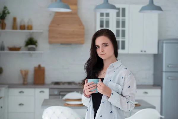 A young girl student stands in the morning in the kitchen with a cup of coffee or tea. Woman dressed in casual clothes jeans and shirt — Stock Photo, Image
