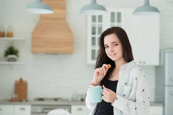 Smiling young brunette woman in the kitchen with a cup of tea and bagels, cookies. Dry snack at lunchtime — Stock Photo, Image