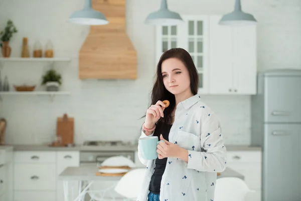 Smiling young brunette woman in the kitchen with a cup of tea and bagels, cookies. Dry snack at lunchtime — Stock Photo, Image