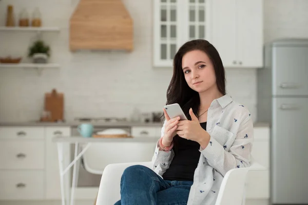 Young brunette girl student is sitting in the kitchen with mobile phone in her hands. Modern gadgets and social networks as a means of communication — Stock Photo, Image