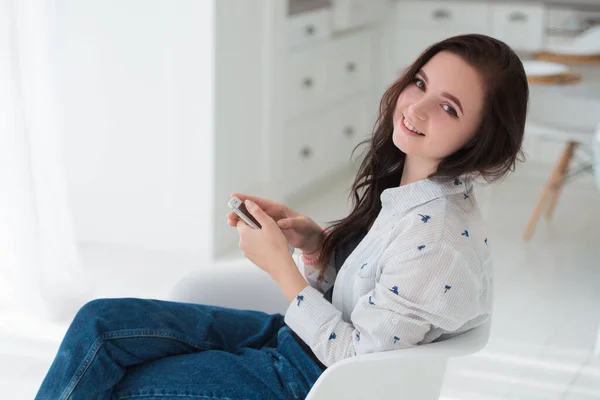 Young brunette woman student is sitting in the kitchen with a mobile smartphone, looking straight and smiling. Modern gadgets or live communication — Stock Photo, Image