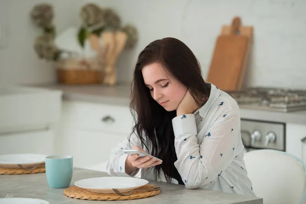 Young brunette girl student sits in the kitchen with mobile smartphone looks at the phone screen. Social networks and communication in modern world — Stock Photo, Image