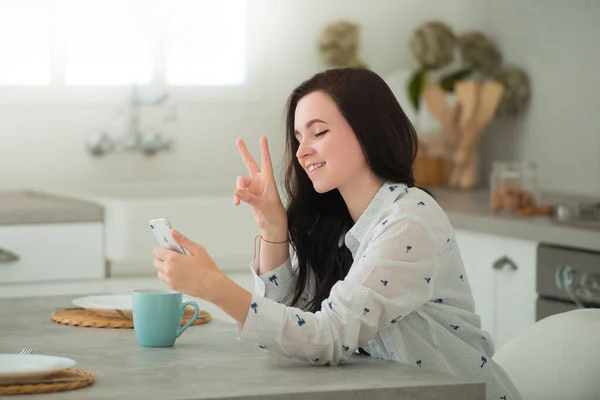 Young brunette woman student is sitting in kitchen with mobile smartphone, smiling and communicating through the screen. Modern gadgets in our life — Stock Photo, Image