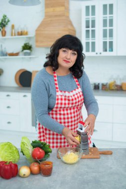 Kareli önlüklü bir kadın pizza malzemesi için mutfakta rendeleyiciye peynir sürüyor..