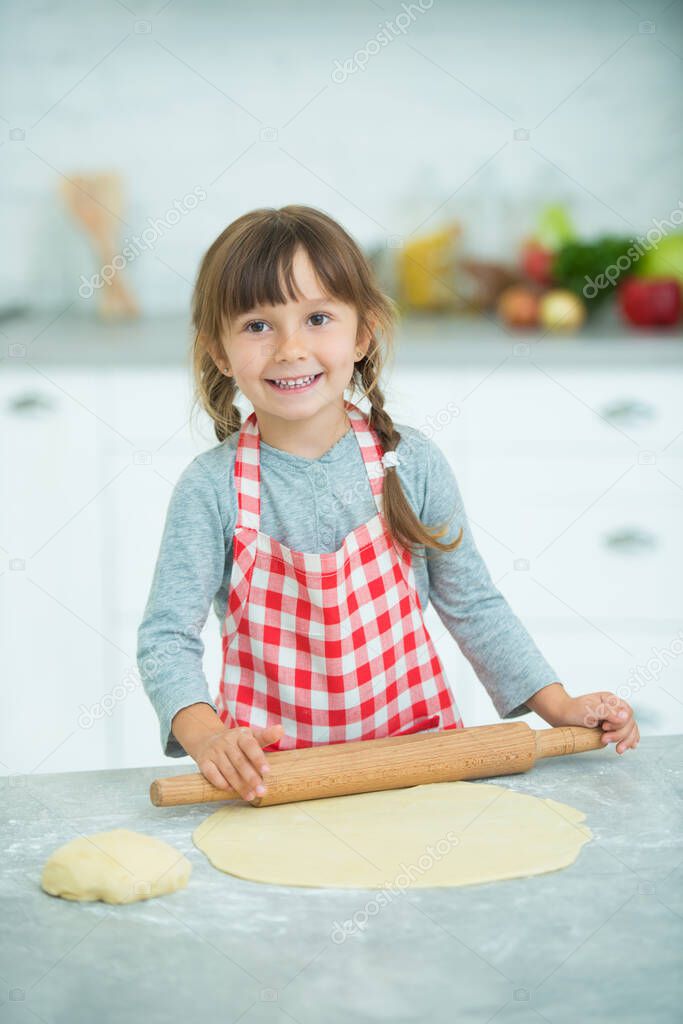 A cute little girl with pigtails in a plaid apron kneads pizza dough on her own. Cooking classes for children.