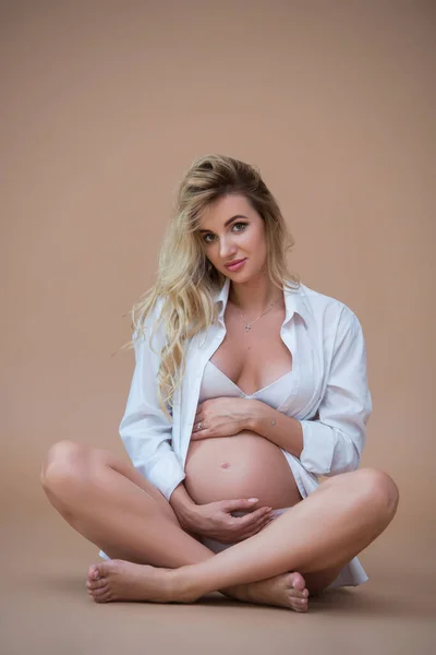美しい妊婦ブロンドの女性はベージュの背景に白いシャツと下着に座って 彼女の手で彼女のおなかを抱擁します 健康な妊娠 — ストック写真