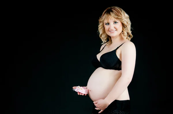 Beautiful pregnant female on black background holding in hand children 's accessory - pink booties — стоковое фото