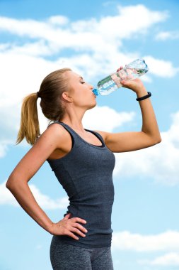 Fitness beautiful blonde woman drinking  water after sports on the background of sky and clouds