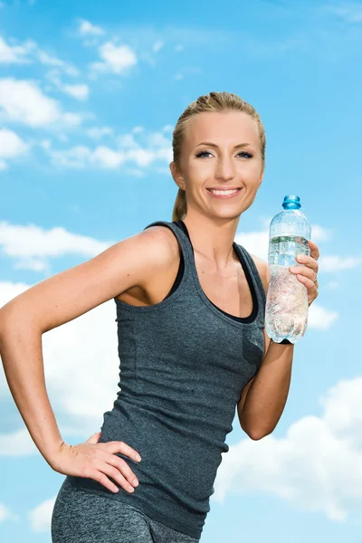 Fitness beautiful blonde woman drinking  water after sports on the background of sky and clouds — Stock Photo, Image