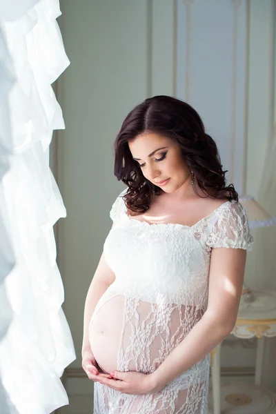 Bela mãe grávida em branco vestido de renda stand na janela e olhar em sua barriga — Fotografia de Stock