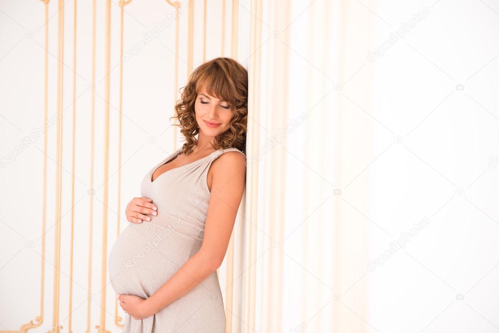 Beautiful red-haired pregnant girl is dreamily leaning against the wall