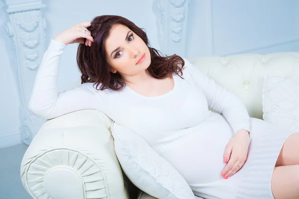 Близкий портрет беременной девушки в белом вязаном платье, сидящей на диване — стоковое фото