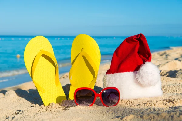 Vacances de Noël en mer. Joyeuses fêtes de nouvel an. Chapeau de Père Noël, sandales, lunettes de soleil sur la plage de sable — Photo