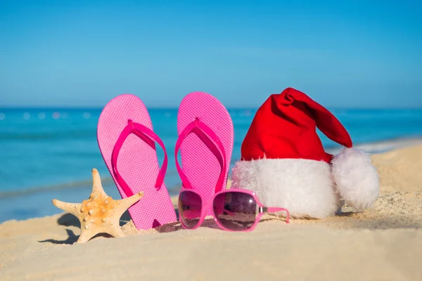 Feliz Año Nuevo y Feliz Navidad en el Mar. Sandalias, gafas de sol y sombrero de santa en la playa de arena . — Foto de Stock