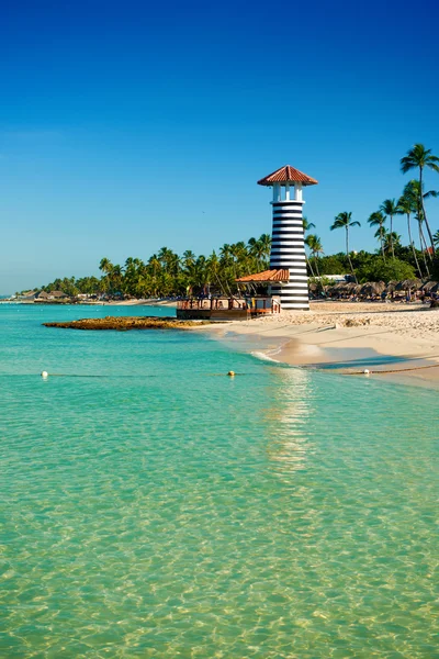 Striped lighthouse on sandy shore with palm trees. Clear water of the Caribbean sea. — Stock Photo, Image