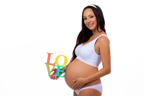 Gravid tjej ler och håller inskriptionen "Love" framför magen. Foto isolerad på en vit bakgrund — Stockfoto