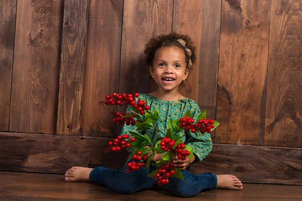 Smiling little african  girl in rural shirt with bunch of berries on  background of brown wooden wall — Stock Photo, Image