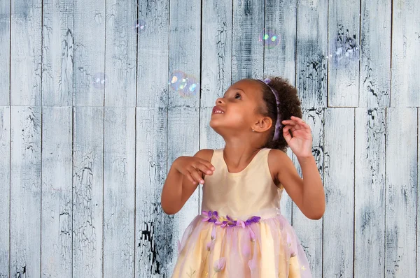 Feliz sorrindo menina africana soprando bolhas de sabão no fundo da parede de madeira — Fotografia de Stock