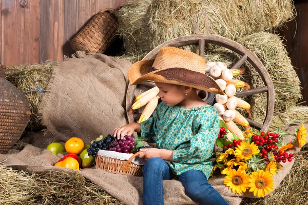 Little african girl in cowboy hat sitting on straw bag with fruits — Stock Photo, Image