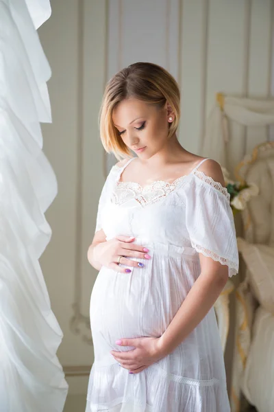 Felicità della maternità. Ragazza incinta con i capelli biondi abbraccia pancia incinta alla finestra — Foto Stock
