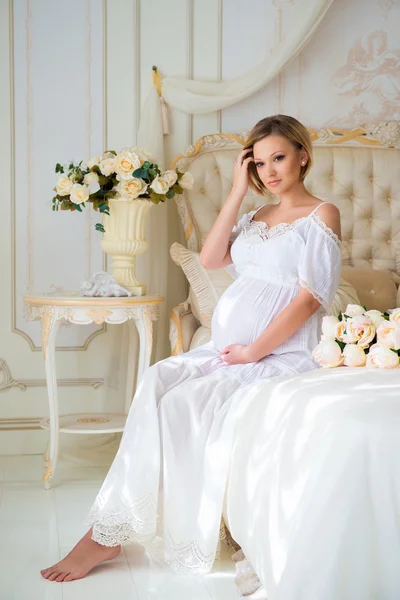 美丽的怀孕女孩在回家的衣服，坐在内部在玫瑰床上 — 图库照片