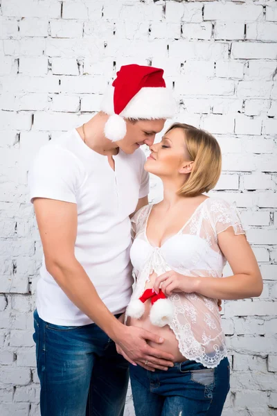 Joven pareja embarazada cerca de una pared de ladrillo blanco con un pequeño niño Santa sombrero — Foto de Stock