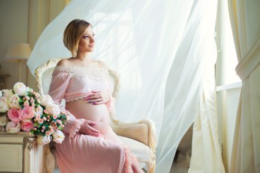 Aşk billowing perdeleri ve hugs göbekle yakın sandalyede oturan sevimli hamile kadın