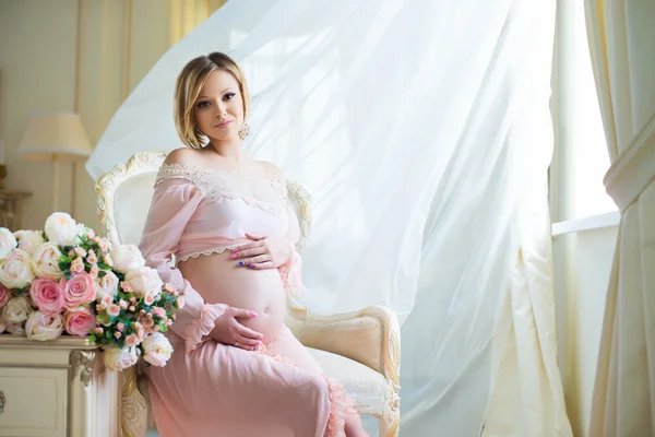 Mulher grávida bonito sentado na cadeira perto de cortinas ondulantes e abraços barriga com amor — Fotografia de Stock