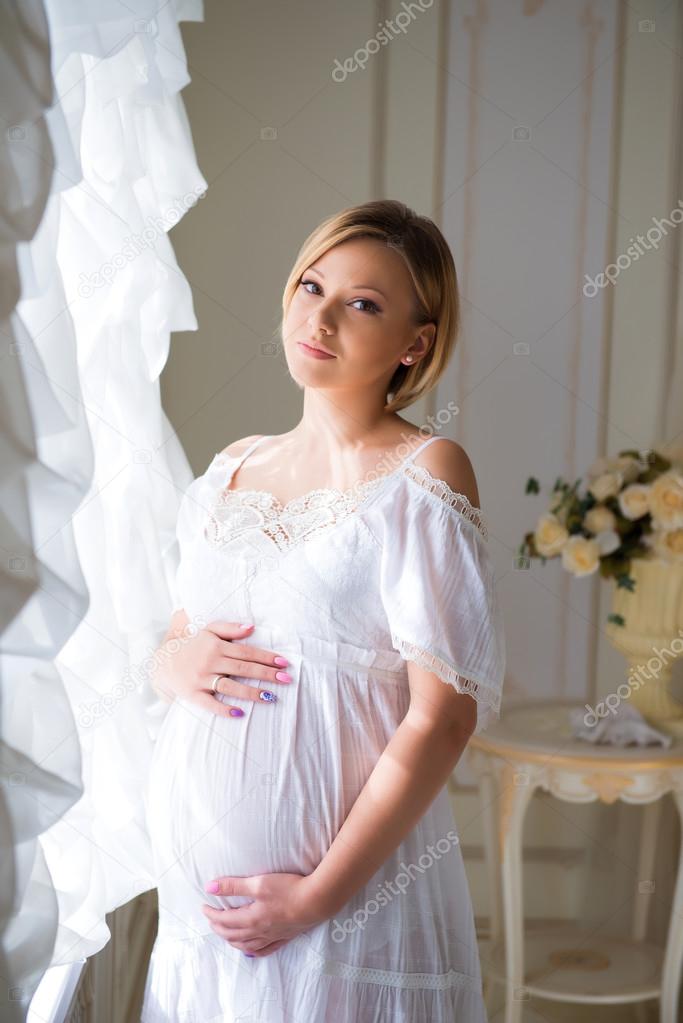 Happiness of motherhood. Pregnant girl with blond hair hugs pregnant belly at the window