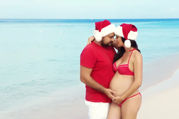 Happy pregnancy, pregnant family. Expectant parents in Christmas costumes and Santa hat on the sea.