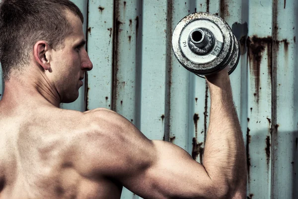Close up young athletic man doing workout with heavy dumbbell against the background of agray metal wall — Stock Photo, Image