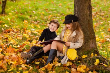 Kız ve oğlan bir ağacın altında oturup, sonbahar güneşli parklarda birlikte kitap okurlar, çocuklar yapraklara oturur, çocuklar kitap okurlar.