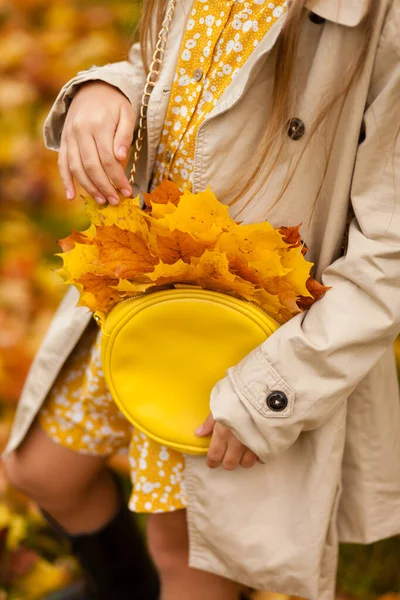 a blonde girl in a beige trench coat and holds a bright yellow handbag with autumn maple leaves, a girl in an autumn park