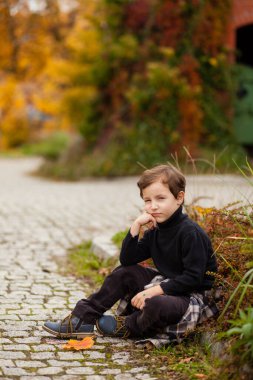 Siyah kazaklı ve kahverengi pantolonlu küçük bir çocuk çalıların yanında oturur, dizlerine yaslanır, sonbaharda parkta yürür.