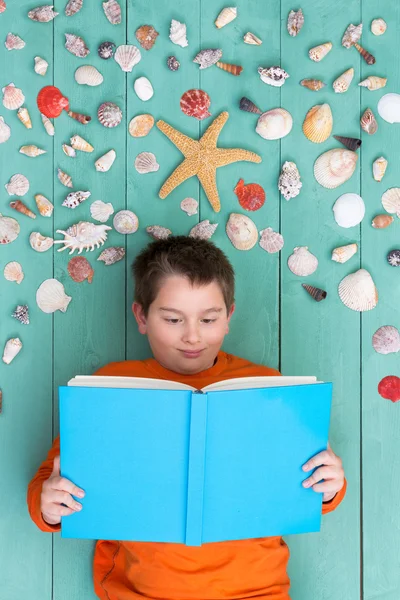 Cute boy reading blank book near seashells — Stock Photo, Image
