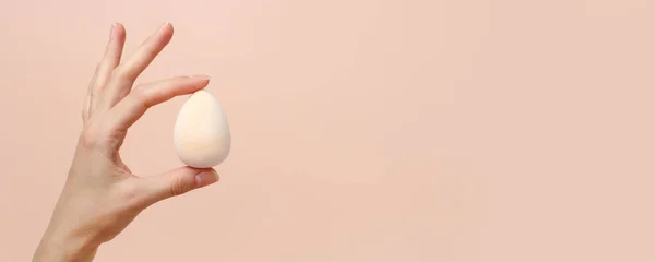 Cosmetic sponge beauty blender in woman hand on a beige background, modern trendy still life, monochrome, earth tones, neutral, beauty industry, makeup artist, face care, copy space, banner