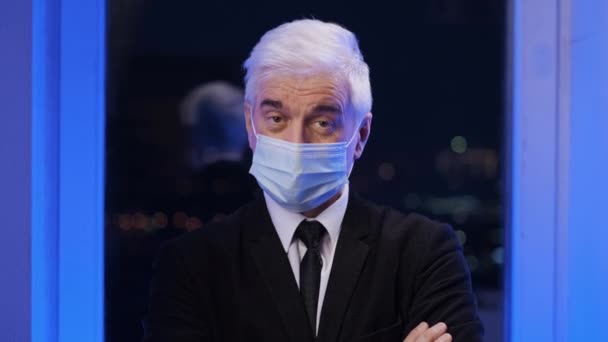 Старший сірий бізнесмен у масці для обличчя дивиться на камеру під портретом коронавірусу . — стокове відео