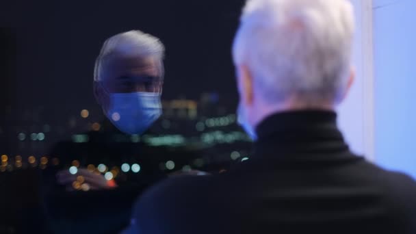 Старик в коронавирусной маске смотрит на свое отражение. Гаффер под пандемией — стоковое видео