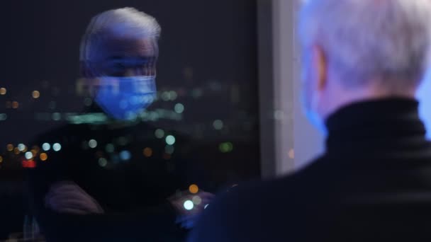 Oudere blanke man in covid-19 epidemie gezichtsmasker kijkt naar reflectie in venster — Stockvideo
