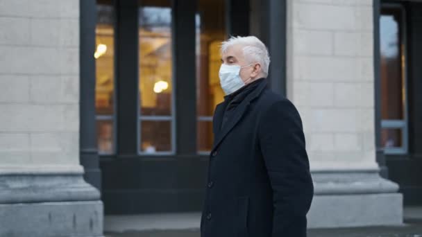 Vieil homme grisonnant en manteau noir se promène près du bâtiment en masque covif en hiver. — Video