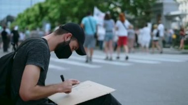 Maskeli isyancı oturmuş pankart çiziyor, işaret levhası ile.