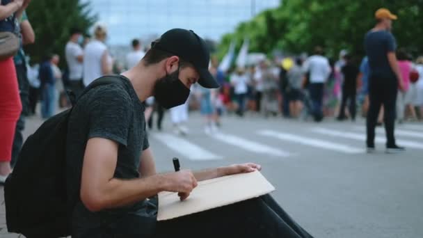 Активист протеста в маске Ковид-19 рисует табличку с надписью "Сидя". Участники митинга. — стоковое видео