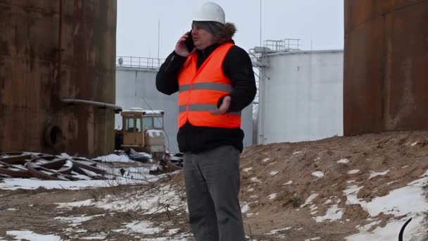Інженер розмовляє на смартфоні на заводі біля нафтових баків взимку — стокове відео