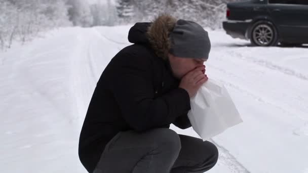 Стресовий чоловік вдихає паперовий мішок біля машини на засніженій дорозі — стокове відео