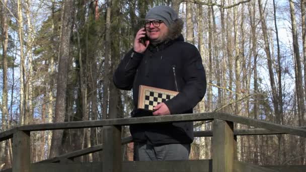 公園の橋の上でスマート フォンで話しているチェスを持つ男 — ストック動画