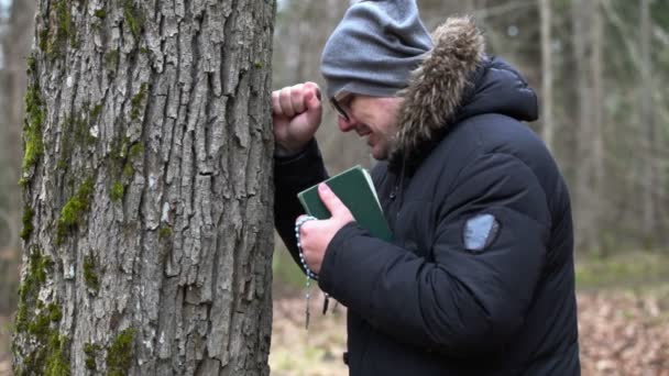 Trauriger Mann mit Bibel und Rosenkranz weint am Baum — Stockvideo